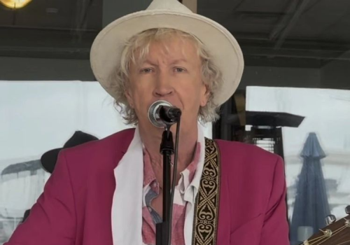 A person in a pink jacket and white hat is playing an acoustic guitar and singing into a microphone.