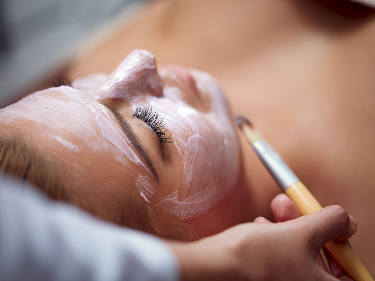 A person is receiving a facial treatment with a white mask applied using a brush, lying down in a spa-like setting.