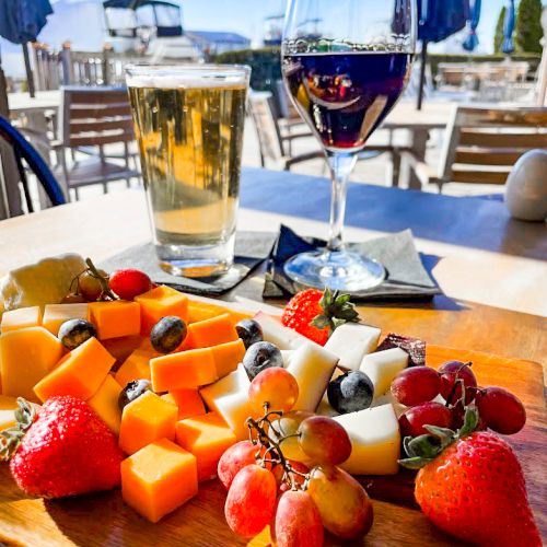 A platter of cheese, strawberries, grapes, and blueberries with a glass of beer and wine on a sunlit outdoor table.