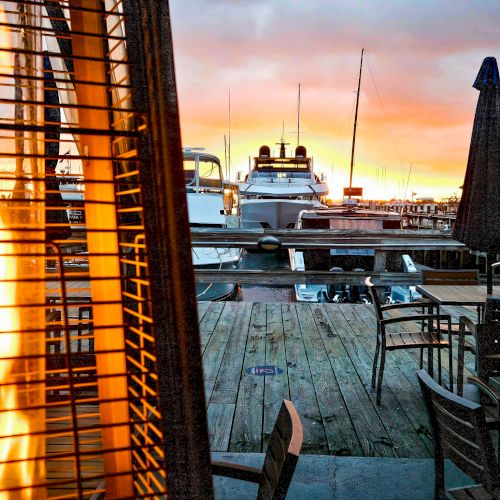 A dockside scene at sunset with boats, outdoor seating, and a lit patio heater creating a cozy atmosphere.