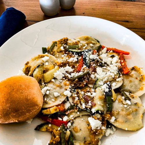 A plate of ravioli with vegetables and cheese, accompanied by a roll. Salt and pepper shakers are in the background.