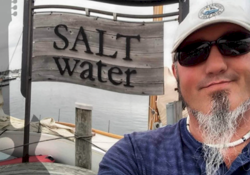 A person wearing sunglasses and a cap poses in front of a "Salt Water" sign near a dock, with boats visible in the background.
