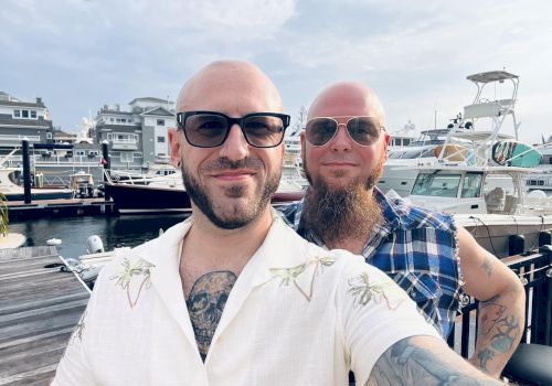 Two bald men with sunglasses posing for a selfie at a marina, boats in the background, one with a tattoo showing on the chest, casual shirts.