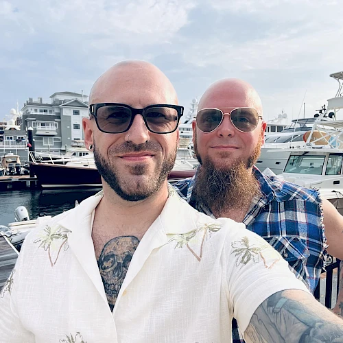 Two bald men with sunglasses posing for a selfie at a marina, boats in the background, one with a tattoo showing on the chest, casual shirts.