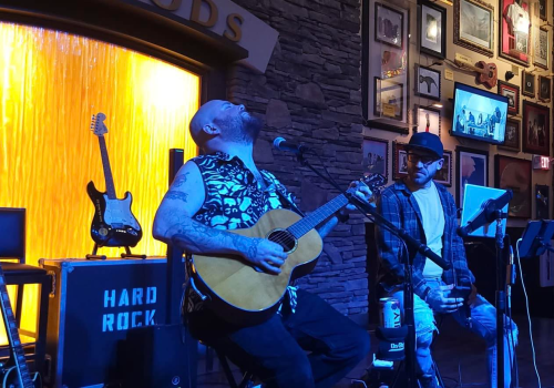 Two musicians performing on stage, one plays guitar while the other sings into a microphone. The setting is a Hard Rock venue.