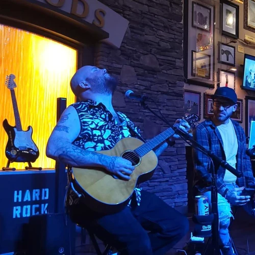 Two musicians performing on stage, one plays guitar while the other sings into a microphone. The setting is a Hard Rock venue.