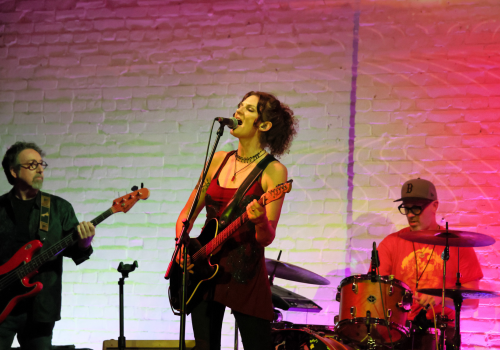 A band performing on stage, with a guitarist singing, a bassist on the left, and a drummer on the right against a colorful lit brick wall.