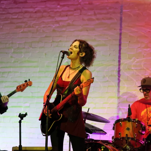 A band performing on stage, with a guitarist singing, a bassist on the left, and a drummer on the right against a colorful lit brick wall.