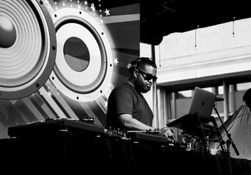 A DJ is performing on stage with audio equipment and a laptop. Large speaker graphics decorate the background in black and white.
