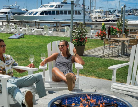Two people relax in Adirondack chairs by a fire pit, with yachts docked in the background and outdoor seating nearby.