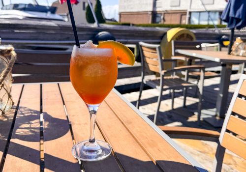 A cocktail with an orange slice on a wooden table, set outdoors with American flags, boats, and buildings in the background.