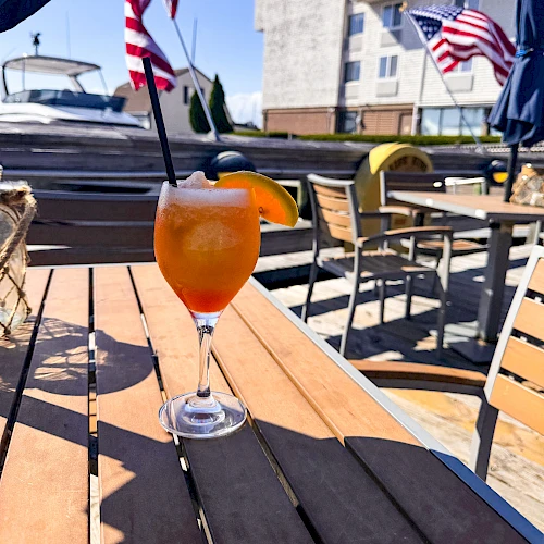 A cocktail with an orange slice on a wooden table, set outdoors with American flags, boats, and buildings in the background.