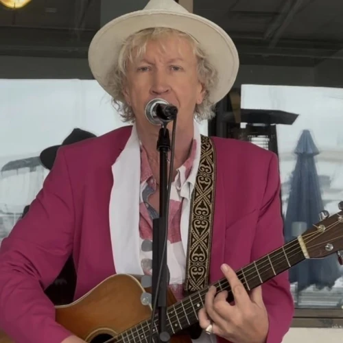 A person in a pink jacket and white hat is playing an acoustic guitar and singing into a microphone.