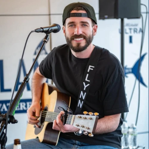 A man is playing an acoustic guitar and singing into a microphone. He is wearing a cap and a "FLYNN" shirt, seated indoors.