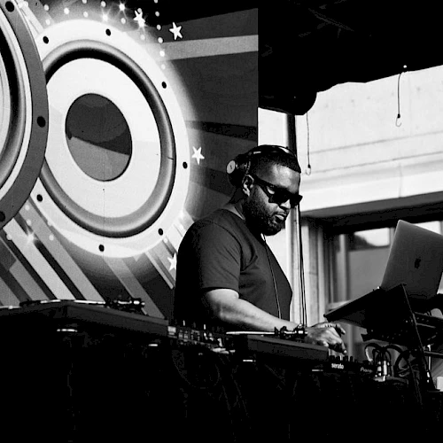 A DJ is performing on stage with audio equipment and a laptop. Large speaker graphics decorate the background in black and white.