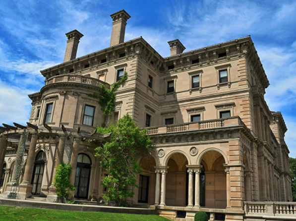 A grand, historic stone mansion with classic architectural details, arches, and greenery against a blue sky.