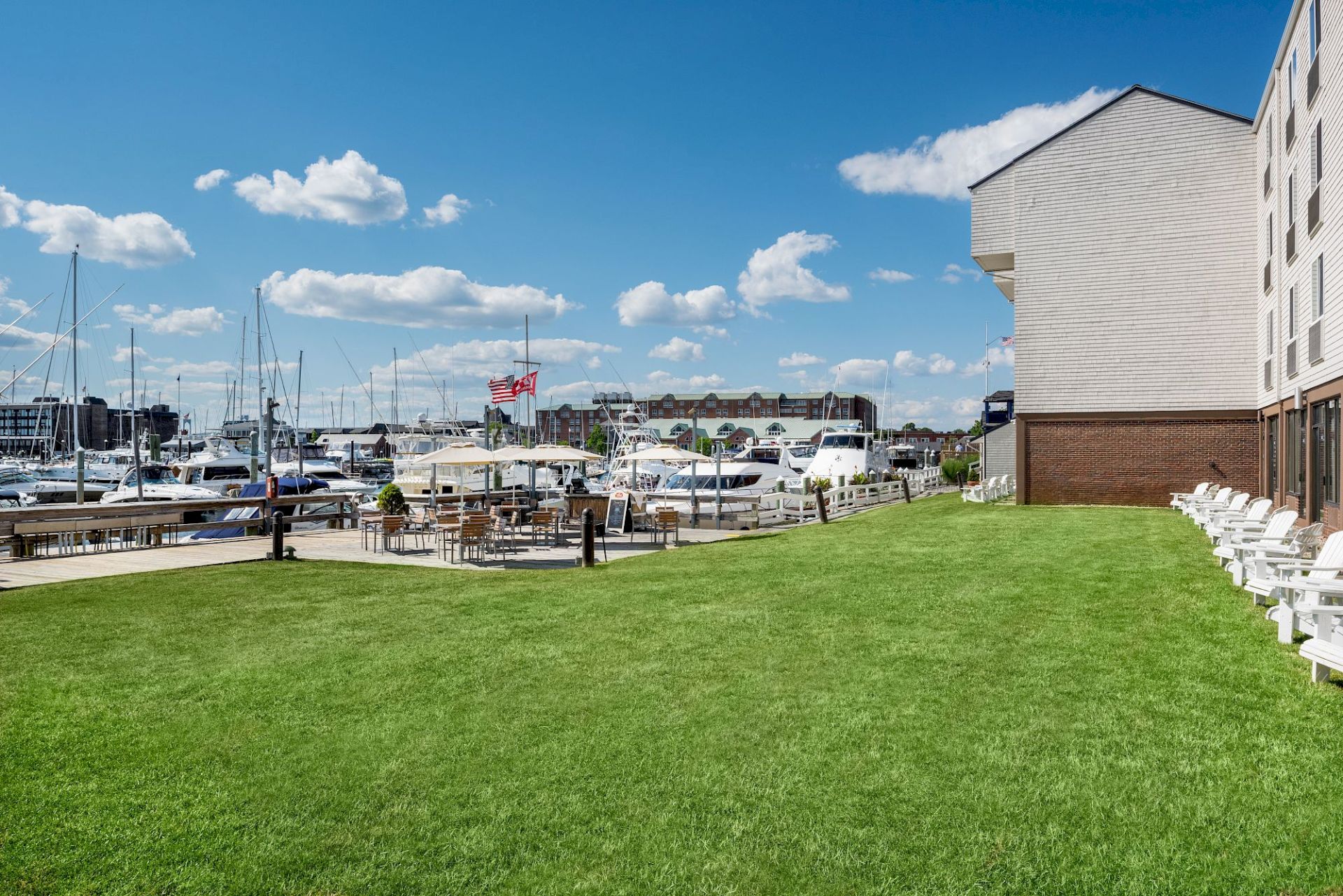 A marina with boats, a grassy lawn, and a white building; sunny with blue skies and fluffy clouds.
