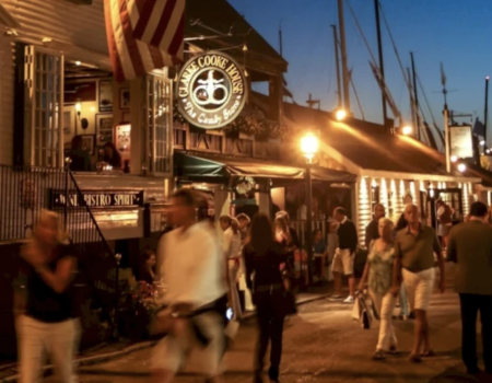 A lively waterfront scene with people strolling near lit shops and boats at dusk, a sign glowing above the crowd, warm lights everywhere.