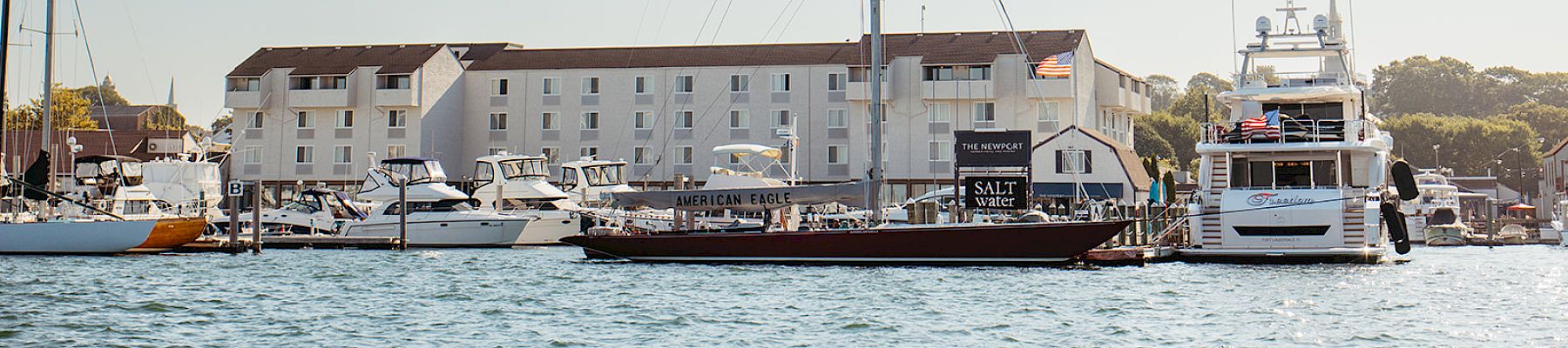 A harbor scene with several boats and yachts docked near a white waterfront building, a tall mast rising among the vessels, calm water.