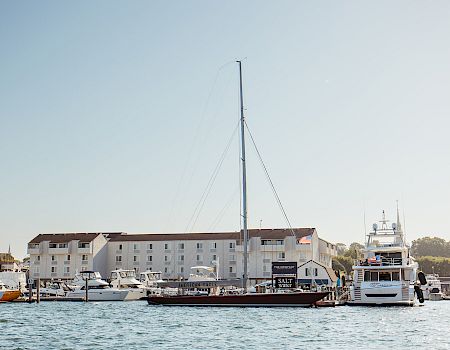 A harbor scene with several boats and yachts docked near a white waterfront building, a tall mast rising among the vessels, calm water.