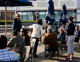 A sunny waterfront restaurant deck with people seated at tables under umbrellas, chatting and enjoying meals by the boats.