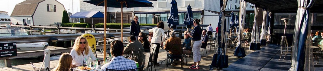 A sunny waterfront restaurant deck with people seated at tables under umbrellas, chatting and enjoying meals by the boats.