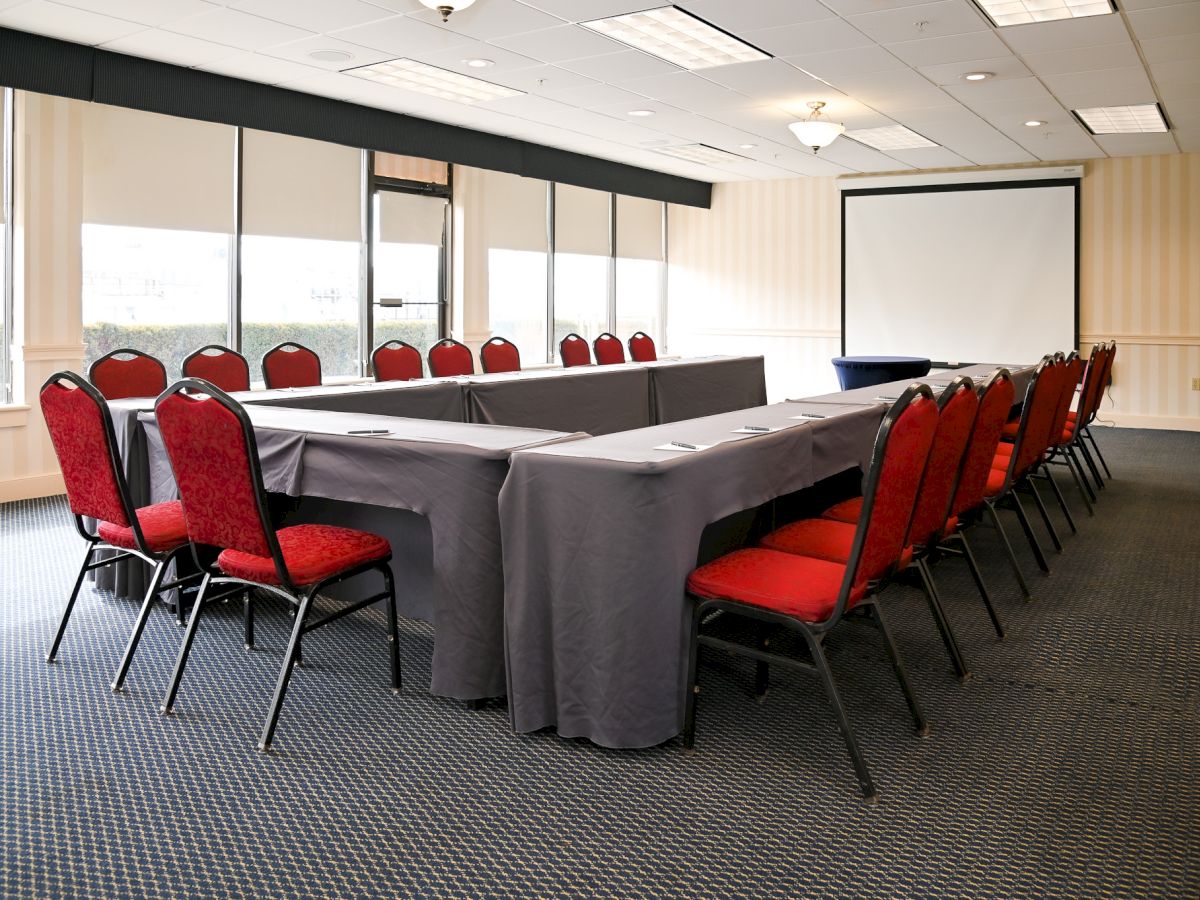 A conference room with a U-shaped table setup, gray tablecloths, red cushioned chairs, large windows, and a projector screen at the front.