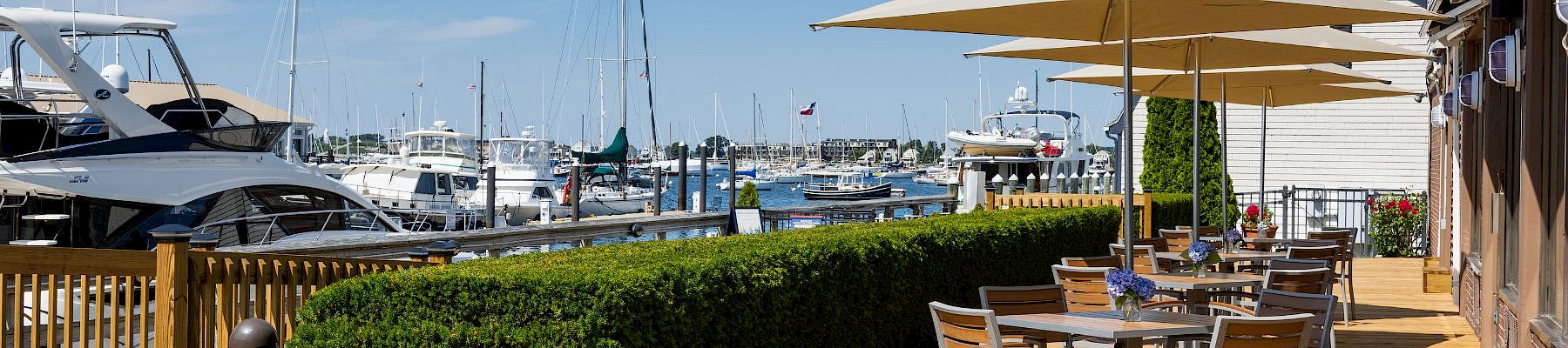 A sunny marina scene with boats, a wooden deck, outdoor seating under umbrellas along the water, and a neatly trimmed hedge.