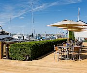 A sunny marina scene with boats, a wooden deck, outdoor seating under umbrellas along the water, and a neatly trimmed hedge.