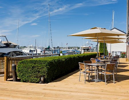 A sunny marina scene with boats, a wooden deck, outdoor seating under umbrellas along the water, and a neatly trimmed hedge.