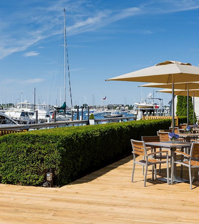 A sunny marina scene with boats, a wooden deck, outdoor seating under umbrellas along the water, and a neatly trimmed hedge.