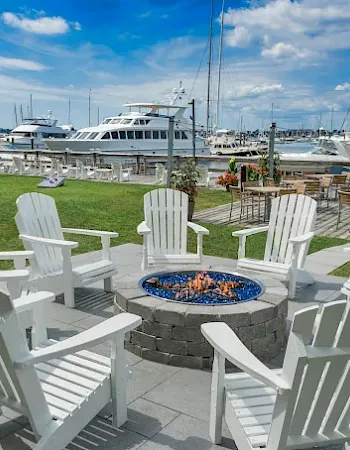 A sunny marina deck with white Adirondack chairs gathered around a round fire pit, with boats docked in the background.