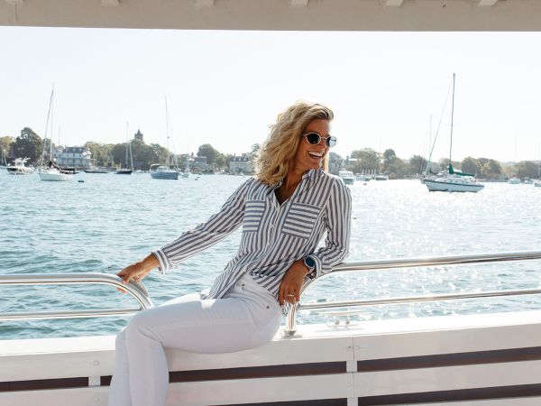 A stylish woman in a striped shirt and white pants sits on a boat railing by a sunny marina, smiling as sailboats drift in the background.