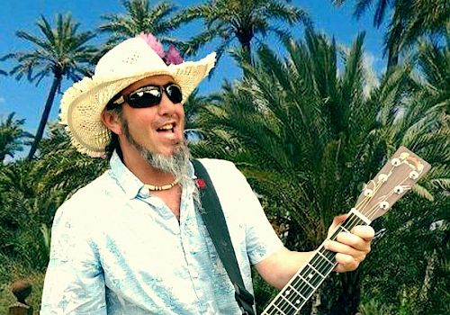 A man with a guitar outdoors, wearing a straw hat and sunglasses, smiling on a wooden path with palm trees in the background.