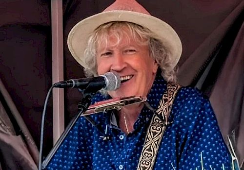 A smiling man playing an acoustic guitar and singing into a microphone on a small stage, wearing a hat and blue polka-dot shirt.