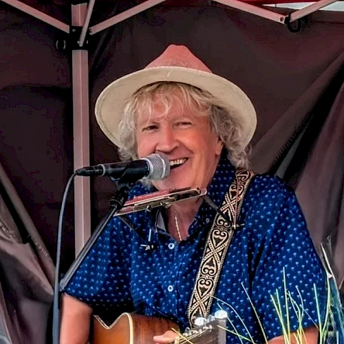A smiling man playing an acoustic guitar and singing into a microphone on a small stage, wearing a hat and blue polka-dot shirt.