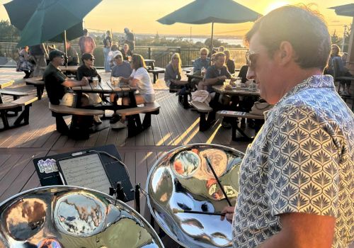 A man plays a shiny steel drum on a sunny outdoor deck with guests dining under umbrellas at sunset, reflections visible in the drums.