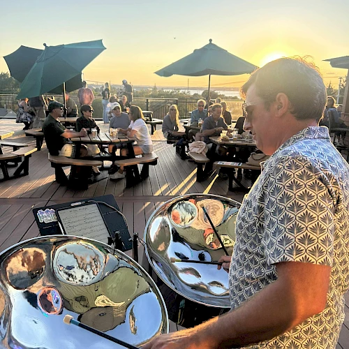 A man plays a shiny steel drum on a sunny outdoor deck with guests dining under umbrellas at sunset, reflections visible in the drums.