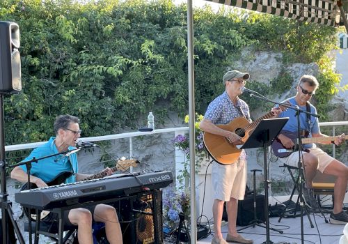 A three-member band performs outdoors: a pianist on left, a youngster singing with a guitar in the middle, and a guitarist seated on the right under a canopy.