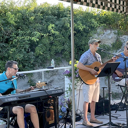 A three-member band performs outdoors: a pianist on left, a youngster singing with a guitar in the middle, and a guitarist seated on the right under a canopy.