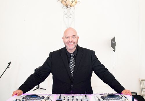 A smiling DJ in a black suit stands behind a glowing DJ console, ready to spin tracks in a bright, minimalist setting.