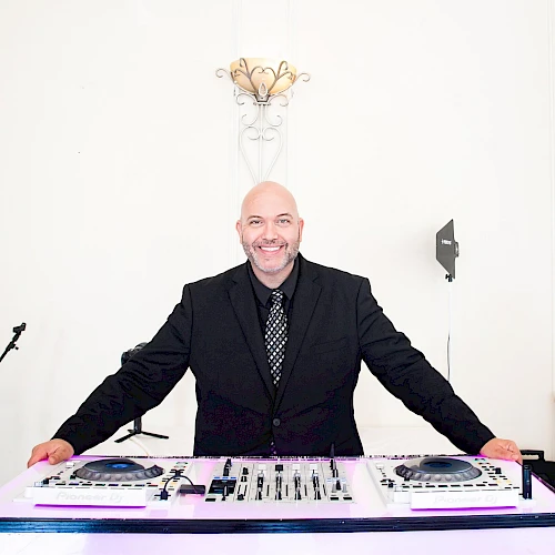 A smiling DJ in a black suit stands behind a glowing DJ console, ready to spin tracks in a bright, minimalist setting.
