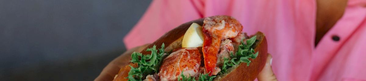 A person in a pink shirt holds a sandwich with leafy greens, lobster or crab meat, and lemon, all inside a crusty roll, delicious.