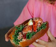 A person in a pink shirt holds a sandwich with leafy greens, lobster or crab meat, and lemon, all inside a crusty roll, delicious.