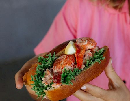 A person in a pink shirt holds a sandwich with leafy greens, lobster or crab meat, and lemon, all inside a crusty roll, delicious.