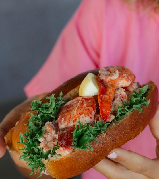 A person in a pink shirt holds a sandwich with leafy greens, lobster or crab meat, and lemon, all inside a crusty roll, delicious.