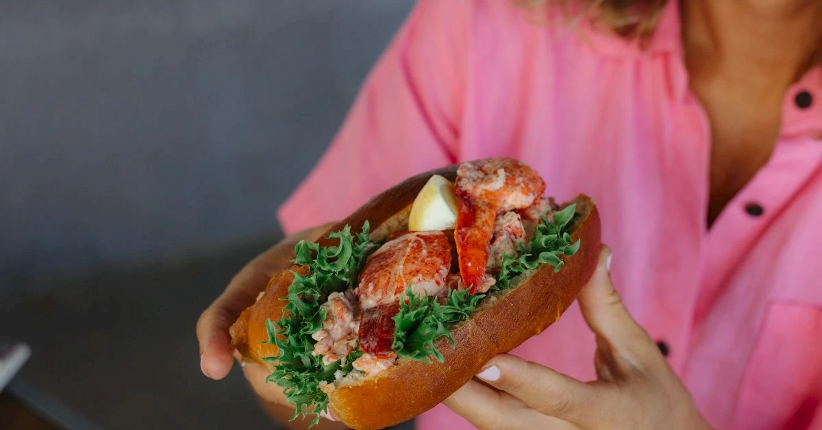 A person in a pink shirt holds a sandwich with leafy greens, lobster or crab meat, and lemon, all inside a crusty roll, delicious.