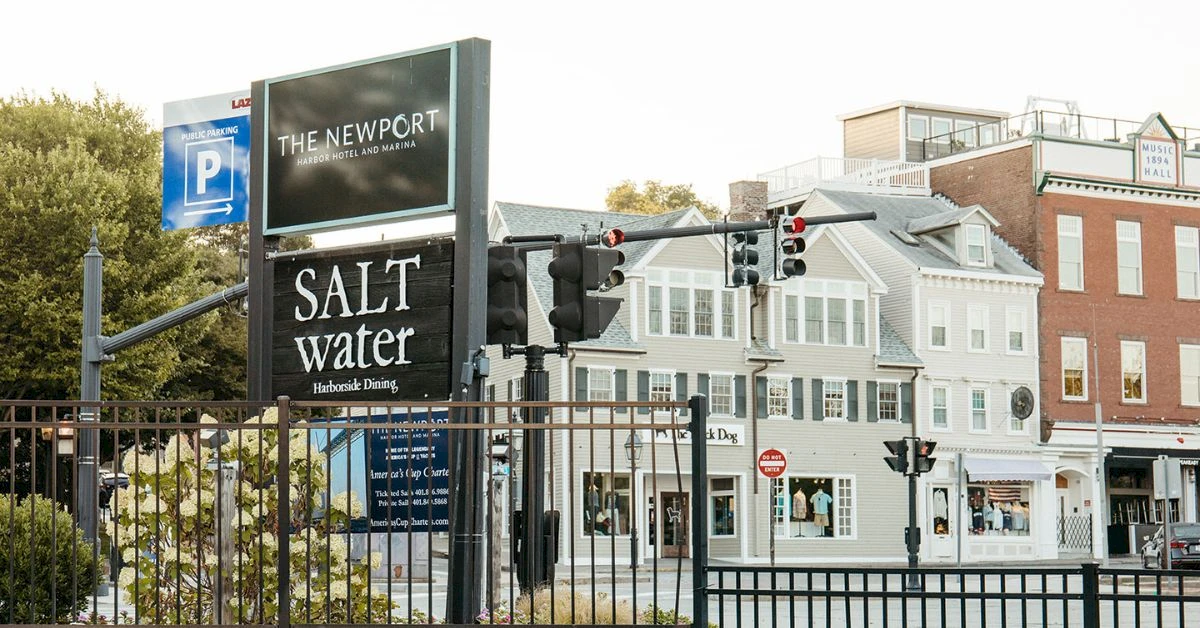 A coastal street scene with a &ldquo;Salt Water&rdquo; sign, traffic lights, a fence, and pastel storefronts in the background. The Newport area appears calm and sunny.