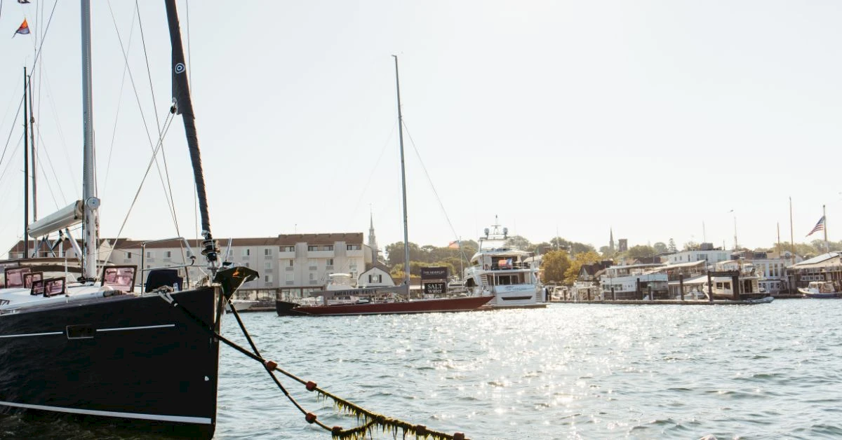 A peaceful harbor scene with sailboats and motorboats docked near a waterfront town; calm water reflects the pale sky, boats lining the quay.