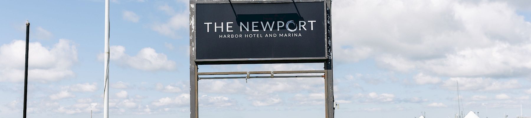A marina pier sign reads &ldquo;The Newport Harbor Hotel and Marina,&rdquo; with blue sky and water in the background.